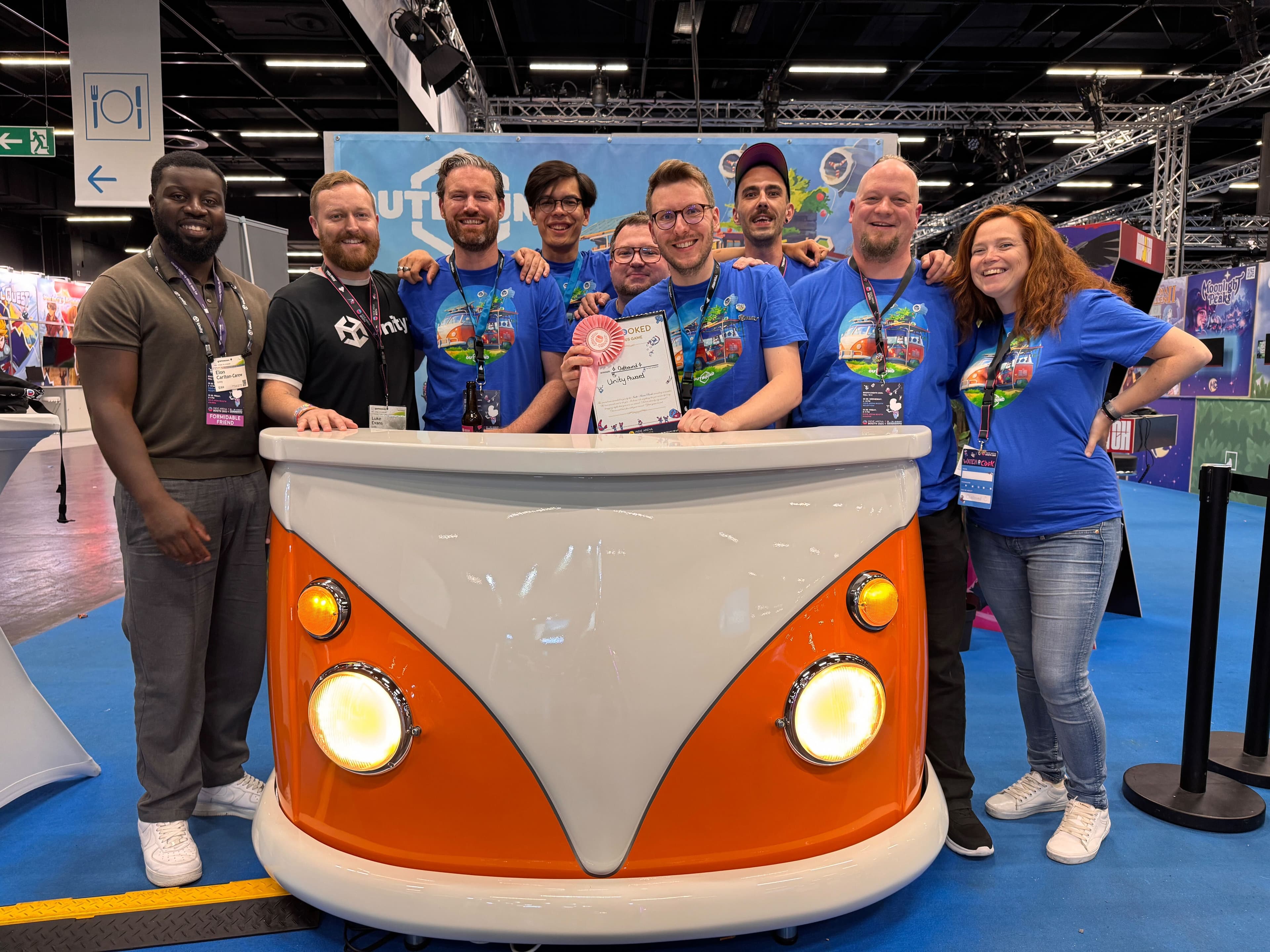 이벤트에서 줄을 서 있는 아홉 사람의 사진입니다. 안경을 쓴 남자가 증명서를 들고 있습니다. 연단은 캠퍼 밴의 후드처럼 보이도록 만들어졌습니다.